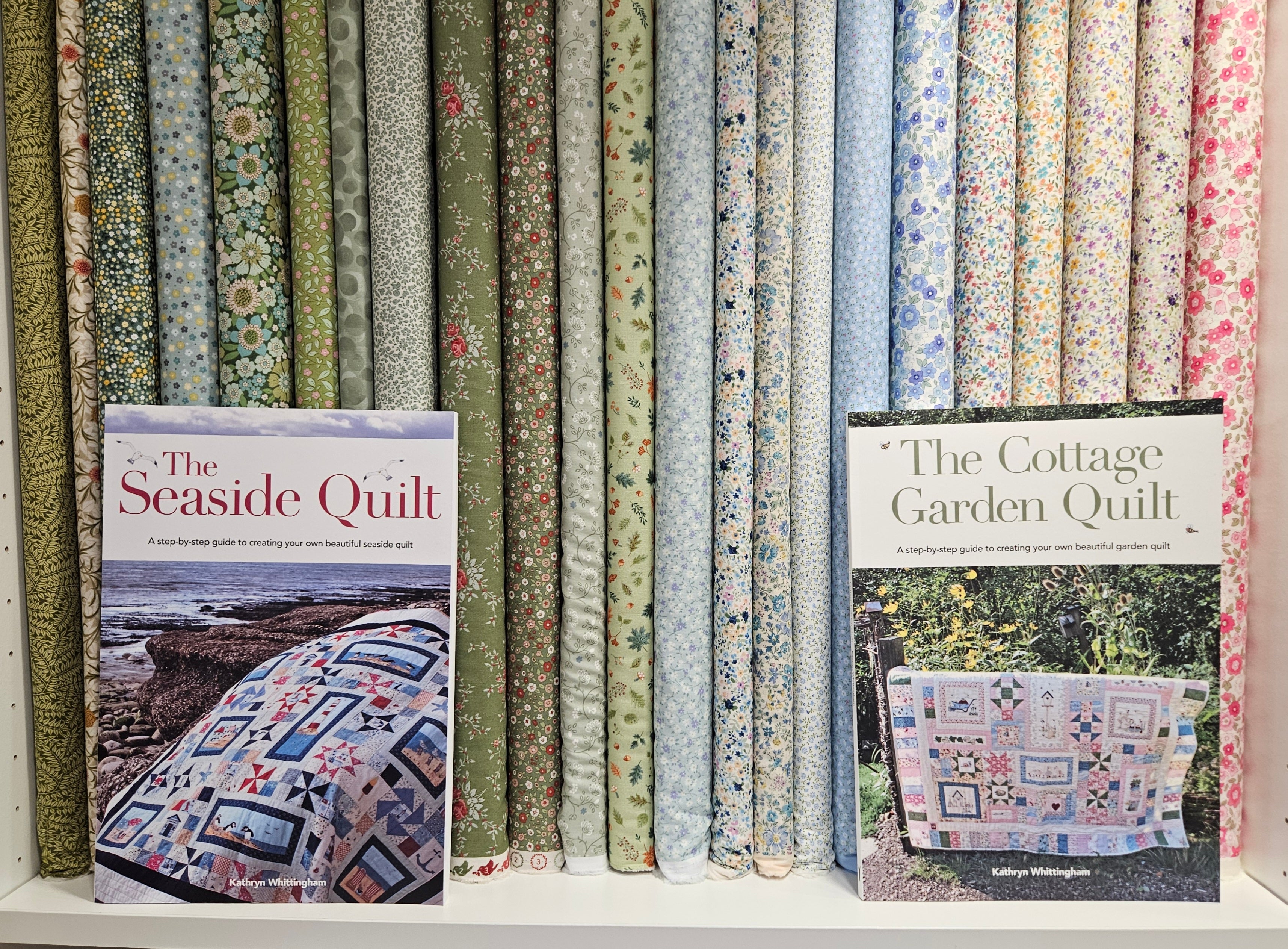 Two books titled 'The Seaside Quilt' and 'The Cottage Garden Quilt' on a shelf with fabric rolls in the background.