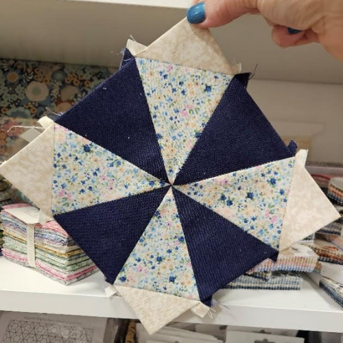 Quilted fabric block with floral patterns held by a hand, with fabric rolls in the background.